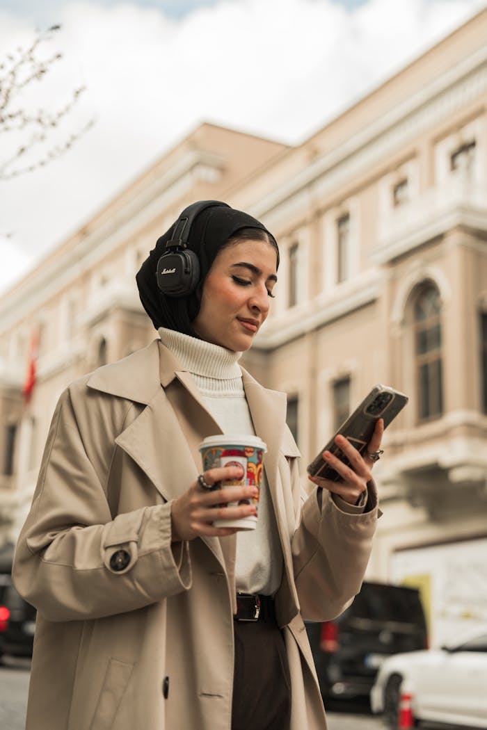 Portfolio Stylish woman in beige coat enjoys coffee and music in city street. Perfect lifestyle moment.