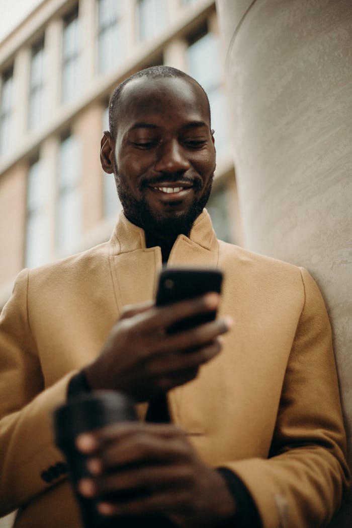 Portfolio A cheerful man in a stylish coat using his smartphone and holding coffee outside.