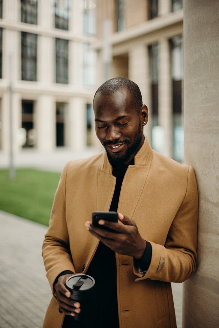 Offerings Relaxed man in coat smiles while using smartphone and holding coffee outside.