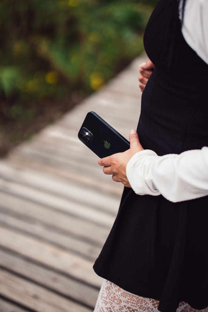 Home Pregnant woman in black dress holds smartphone, walking on wooden path outdoors.