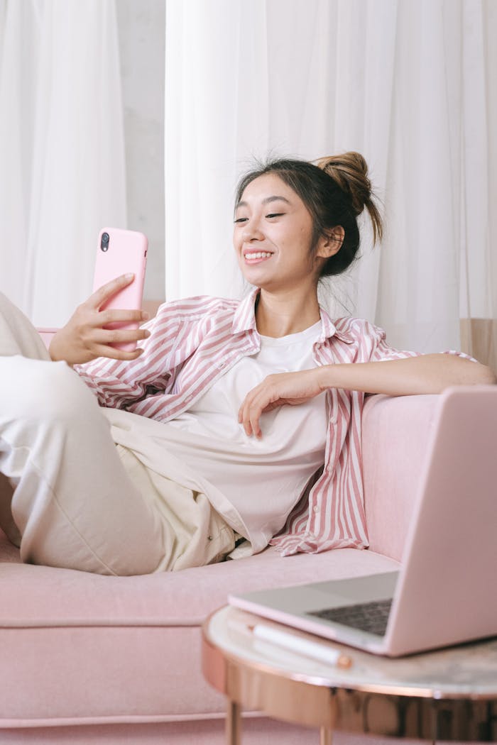Offerings Asian woman relaxing on a pink sofa, using a smartphone and smiling at home.