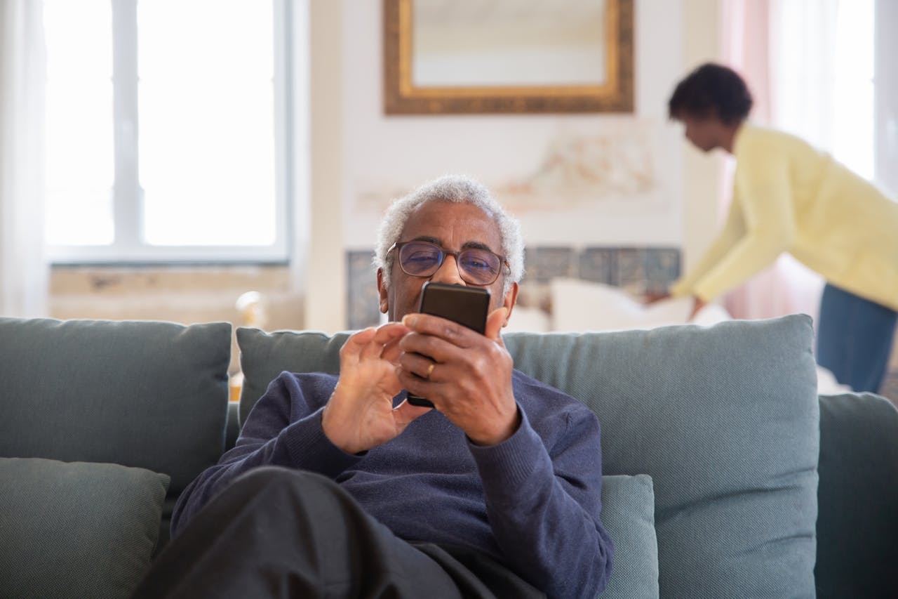 Offerings Elderly man sitting on couch using smartphone indoors. Cozy home setting.